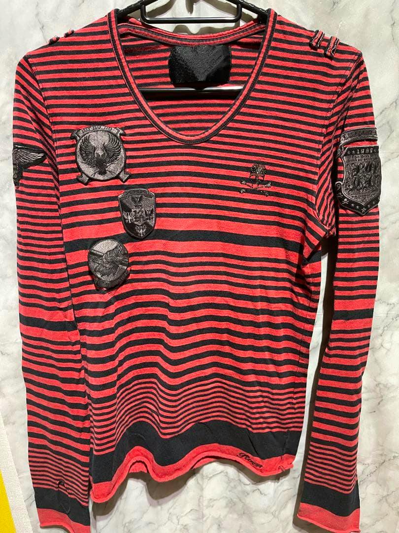 Red and black striped long-sleeve Roen top with black embroidered skull and crest patches hanging on a hanger.