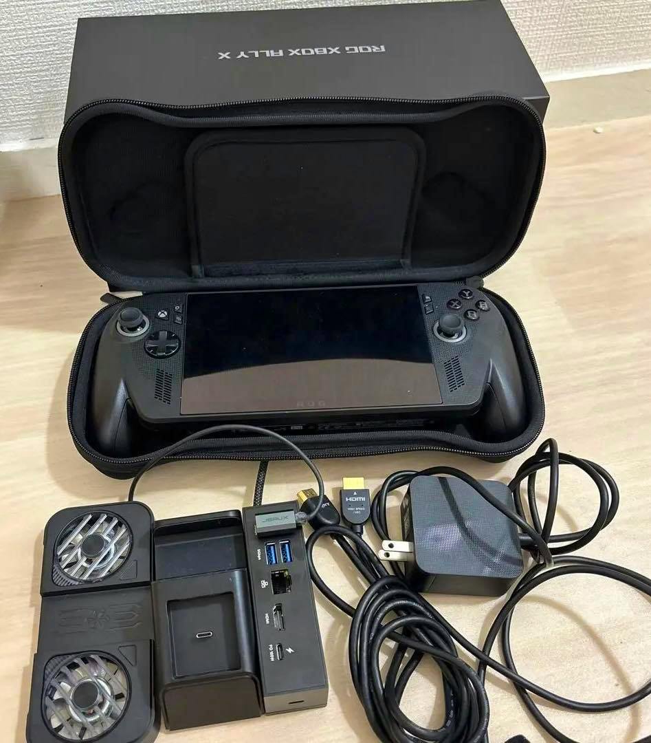 ROG Xbox Ally X handheld console shown inside an open carrying case with its dock, cooling fans, and cables arranged in front.
