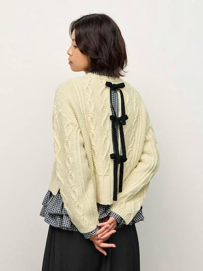 Model wearing a cream cable-knit sweater showing three black ribbon bows down the back.