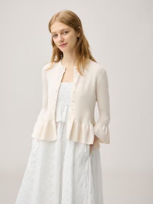 A model wearing a cream-colored cardigan with a peplum hem and button front over a white dress.
