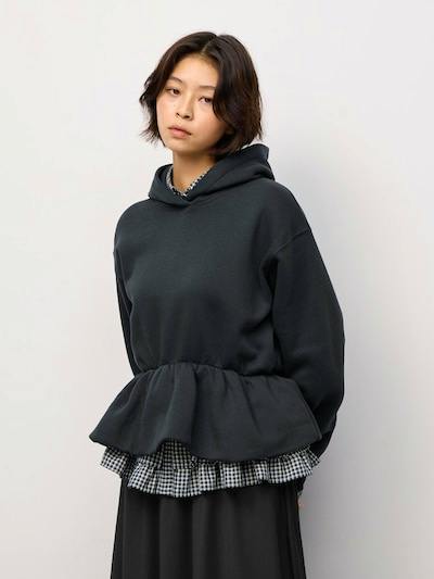 Model wearing a dark hooded sweatshirt with a ruffled peplum hem over a checked shirt.