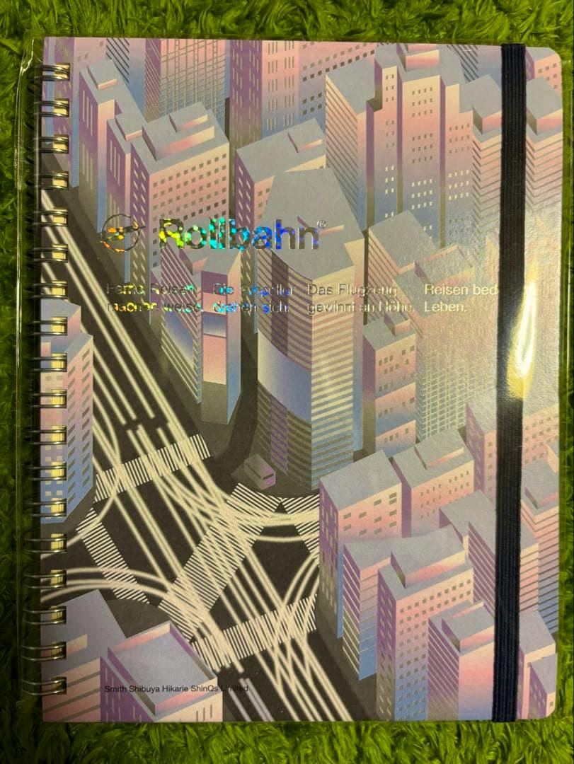 Front view of a Rollbahn spiral-bound notebook with a pastel cityscape cover and a black elastic band, photographed on a green surface.