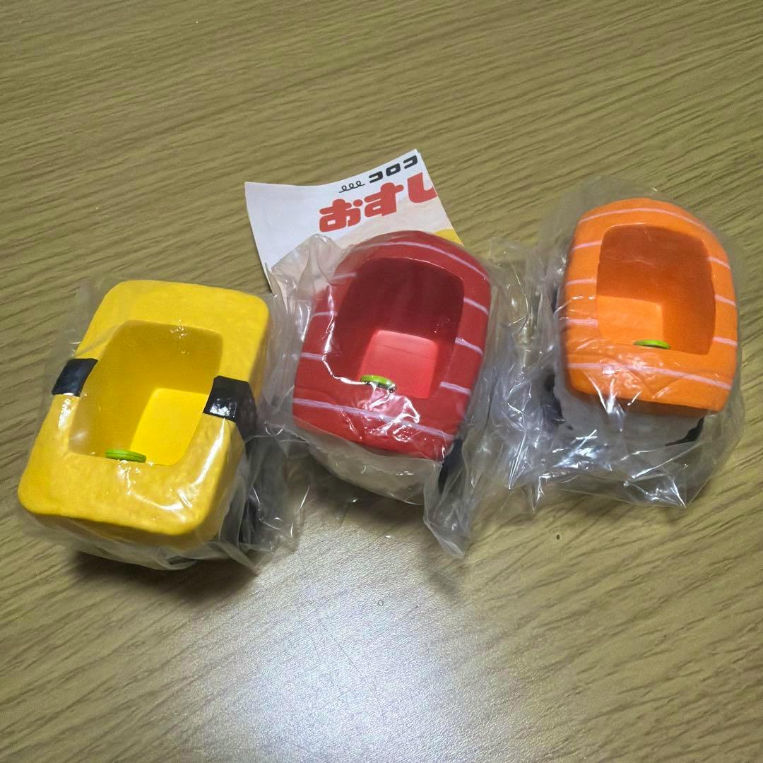 Three plastic sushi-themed toy cars (yellow, red, orange) wrapped in clear plastic on a wooden table.
