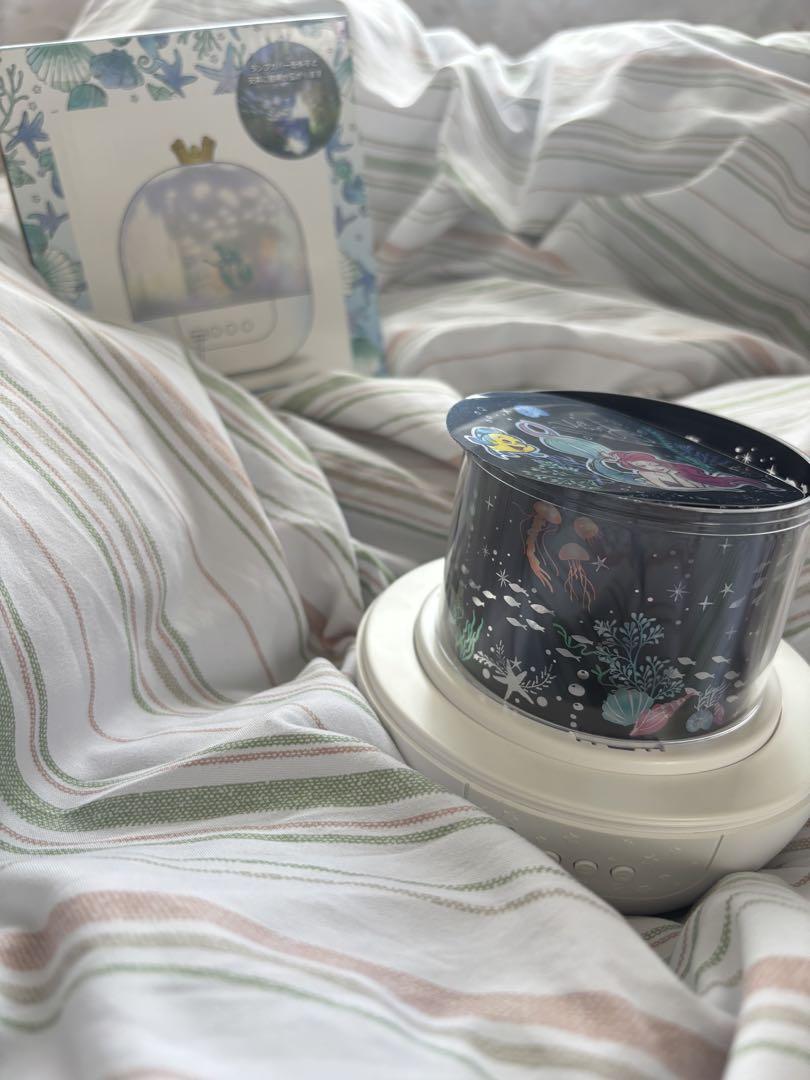 A small room projector with a mermaid-themed decorative cylinder on a white base, photographed on striped bedding with its box in the background.