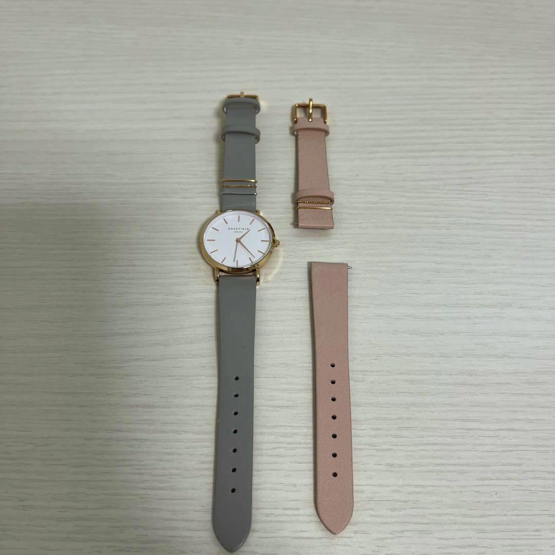 A ROSEFIELD wristwatch with a gray strap and gold case laid flat next to two pink replacement strap pieces on a light wood surface.