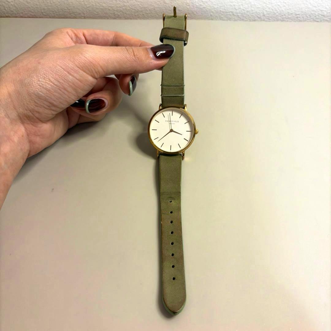 A ROSEFIELD gold-cased wristwatch with a green leather strap held by a hand on a light surface.