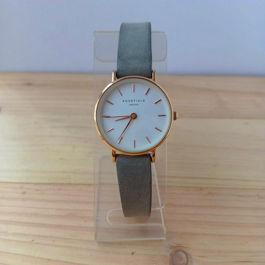 Front view of a ROSEFIELD quartz wristwatch with a white dial, rose‑gold case and grey strap displayed on a clear stand.