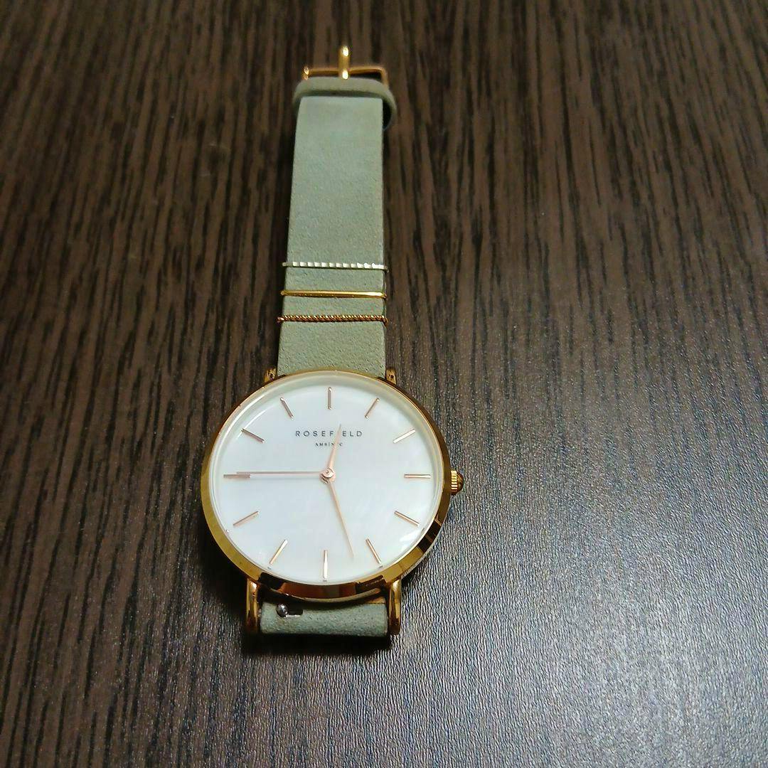 ROSEFIELD wristwatch with a light green strap and gold-tone case placed on a dark wood surface.