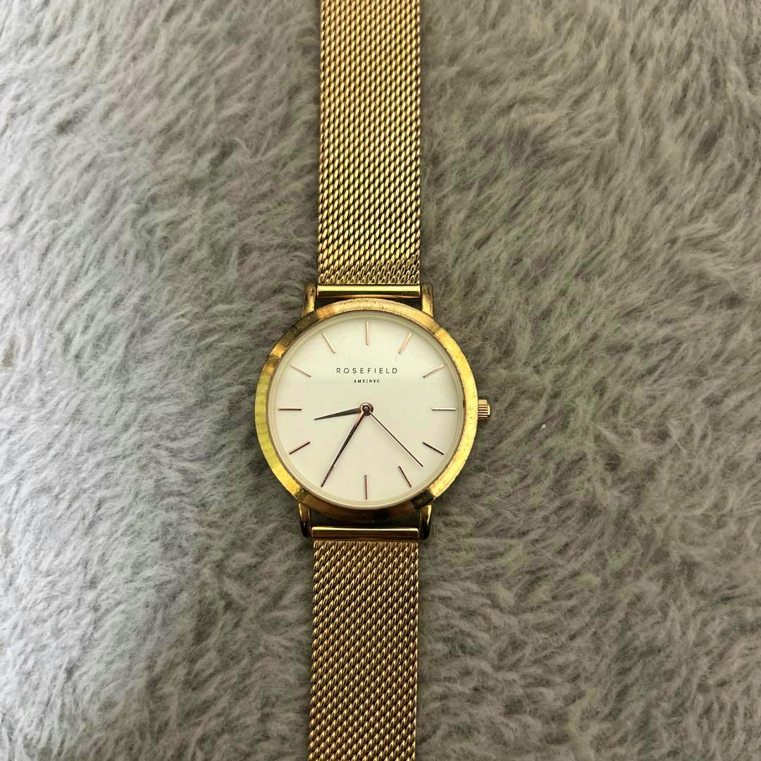 Close-up of a gold Rosefield women’s wristwatch with a white dial and gold mesh band on a gray fuzzy background.