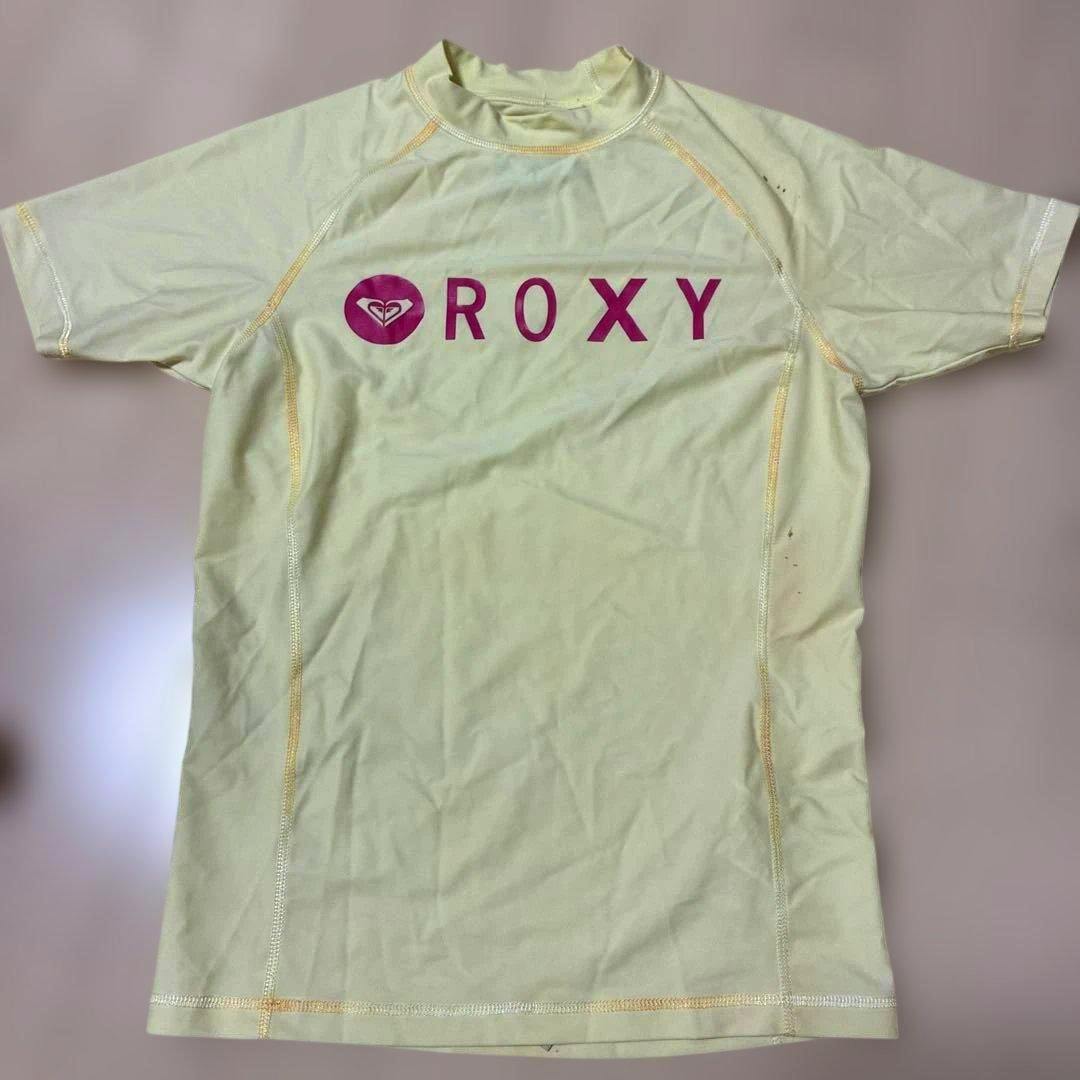 Front view of a light yellow ROXY rash guard with a pink ROXY logo laid flat.