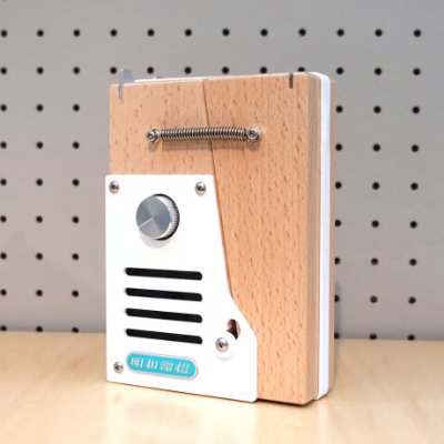 A beech compact speaker showing its white metal front grille, volume knob, and wooden body against a pegboard background.