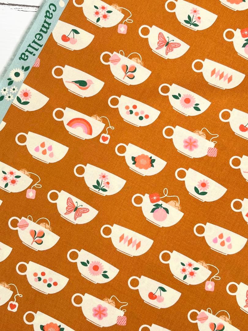 Brown cotton fabric printed with repeating cream teacup motifs decorated with small floral, fruit, and butterfly illustrations.