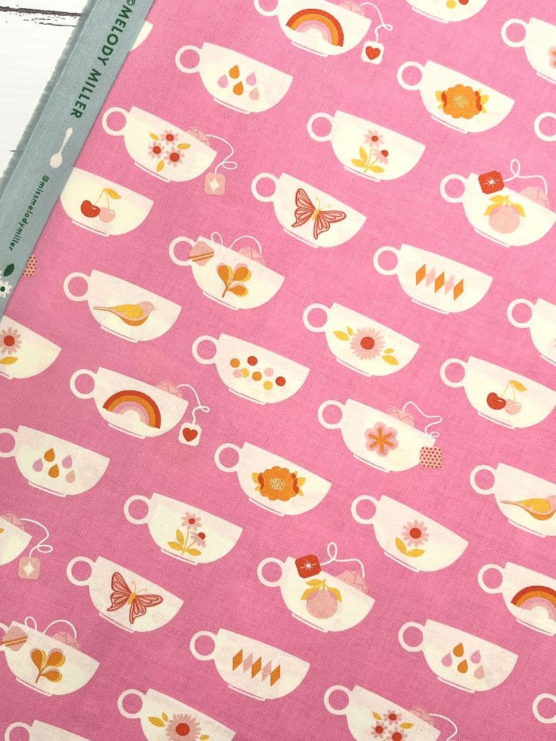 Pink dyed cotton fabric showing repeating white coffee-cup motifs with small floral, rainbow, and fruit decorations.