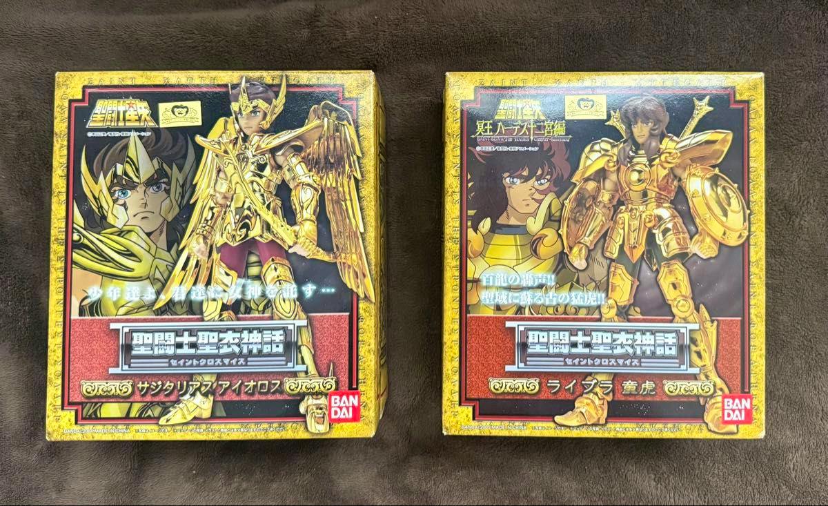 Two boxed Bandai Saint Cloth Myth figures photographed on a dark fabric background, Sagittarius Aiolos box at left and Libra (童虎) box at right.