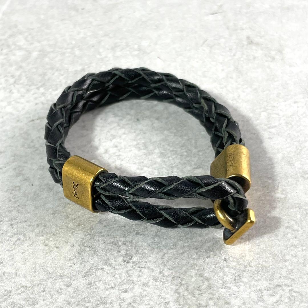 Black braided cow leather bracelet with gold-tone YSL logo hardware shown on a light surface.