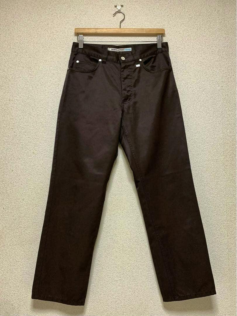 Front view of SAINT LAURENT JEANS men's cotton pants hanging on a hanger against a wall.