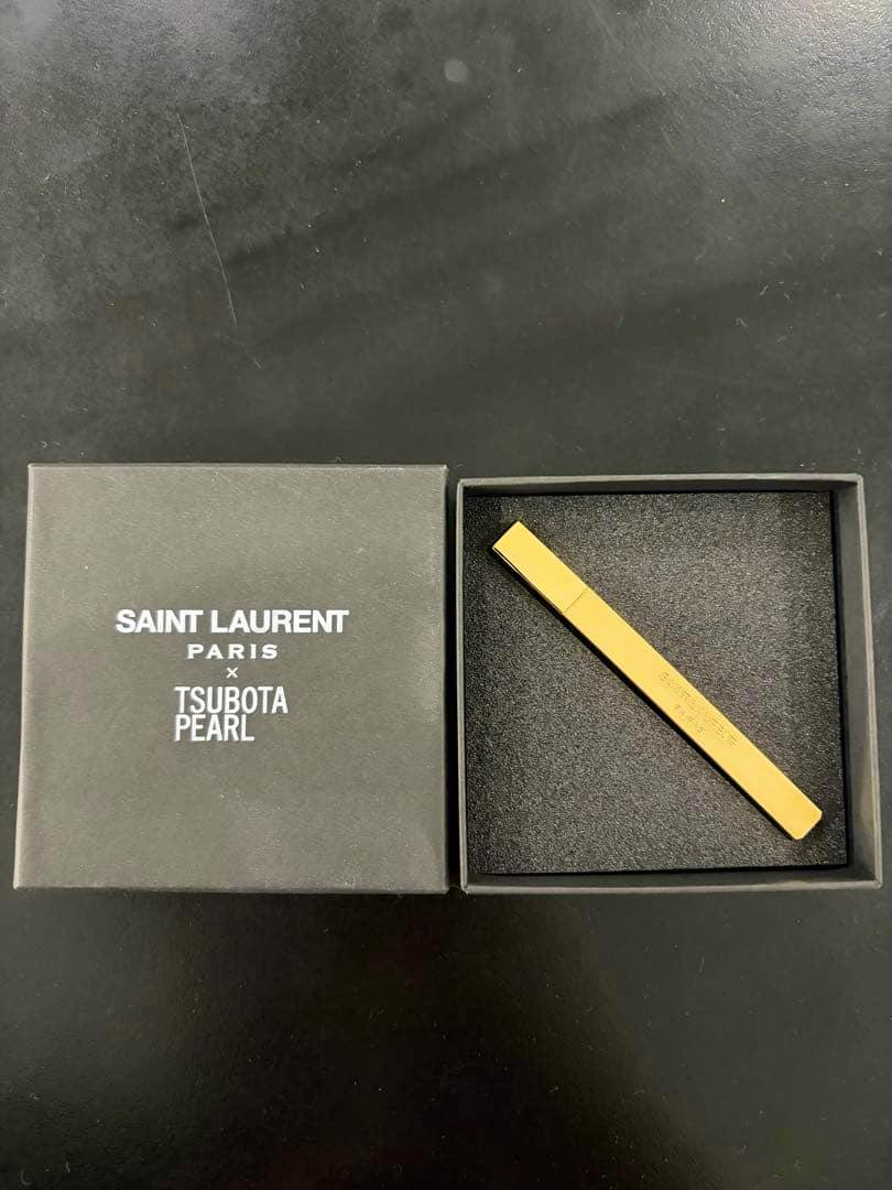 Gold rectangular Saint Laurent × Tsubota Pearl lighter case displayed in a foam-lined box beside the branded box lid.