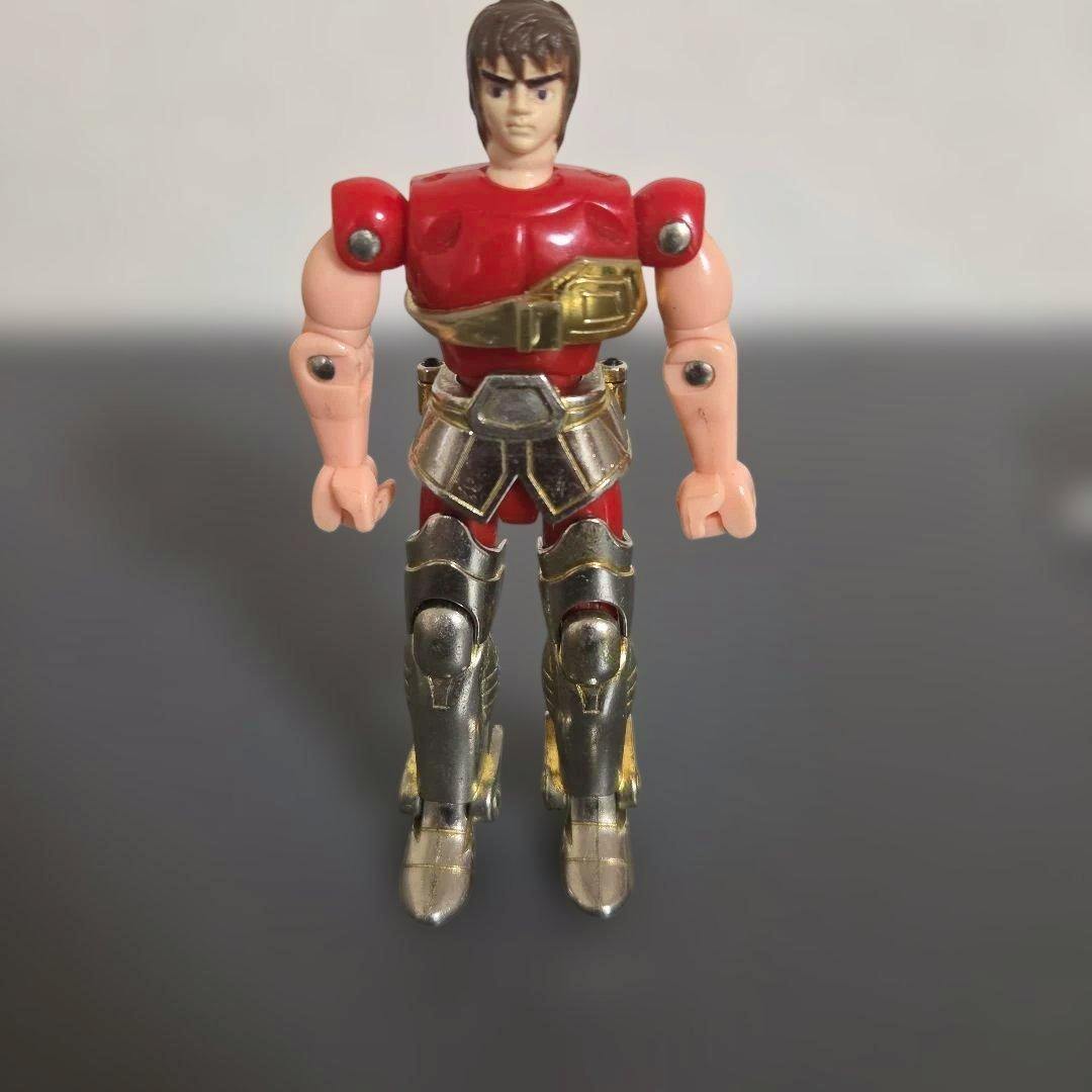 Front view of the Saint Seiya plastic figure wearing red and metallic armor with gold accents, standing on a gray surface.