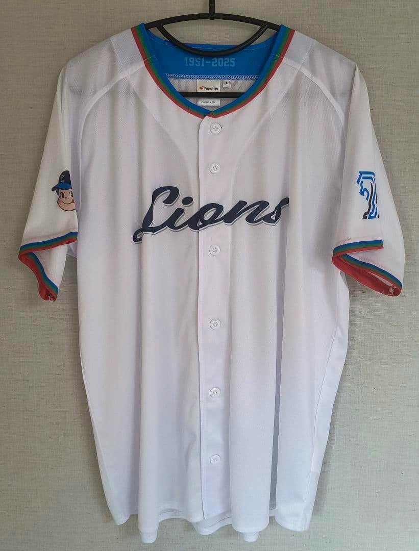 Front view of the white Seibu Lions polyester uniform (size L) with "Lions" script across the chest, button-front, and a Peko-chan graphic on the sleeve, shown hanging on a hanger.