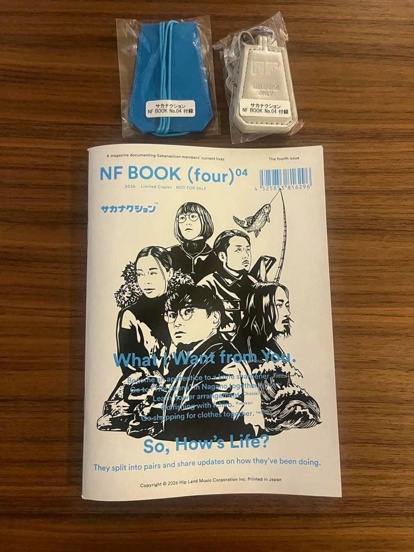 Photograph of the サカナクション NF BOOK (four)04 magazine on a wooden surface with two sealed bonus accessory items placed above it.