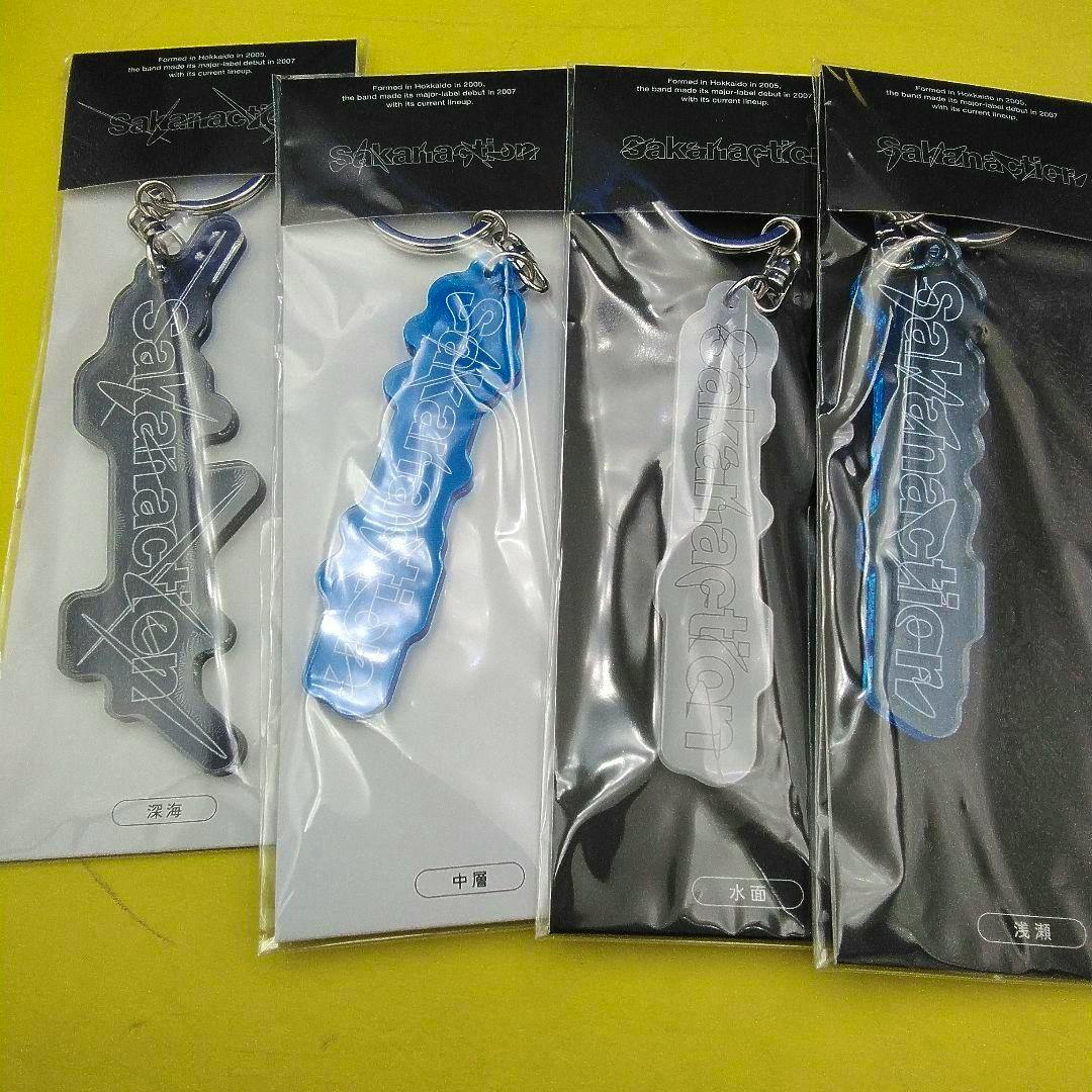 Four packaged Sakanaction STATUS LOGO acrylic keychains displayed side-by-side on a yellow background.