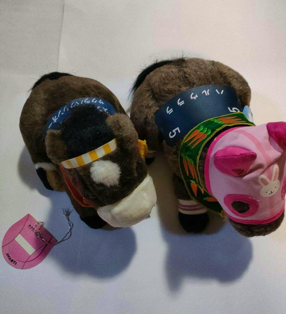 Top-down photo of two brown horse plush toys (競馬ぬいぐるみ) with decorative headbands and one wearing a pink hood on a white background.