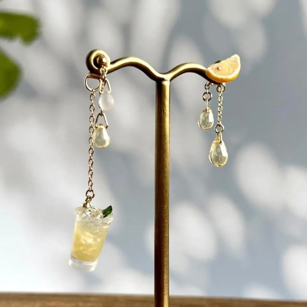 A pair of salty dog-themed earrings on a stand, one with a small drink charm and the other with a lemon-slice charm and teardrop beads.