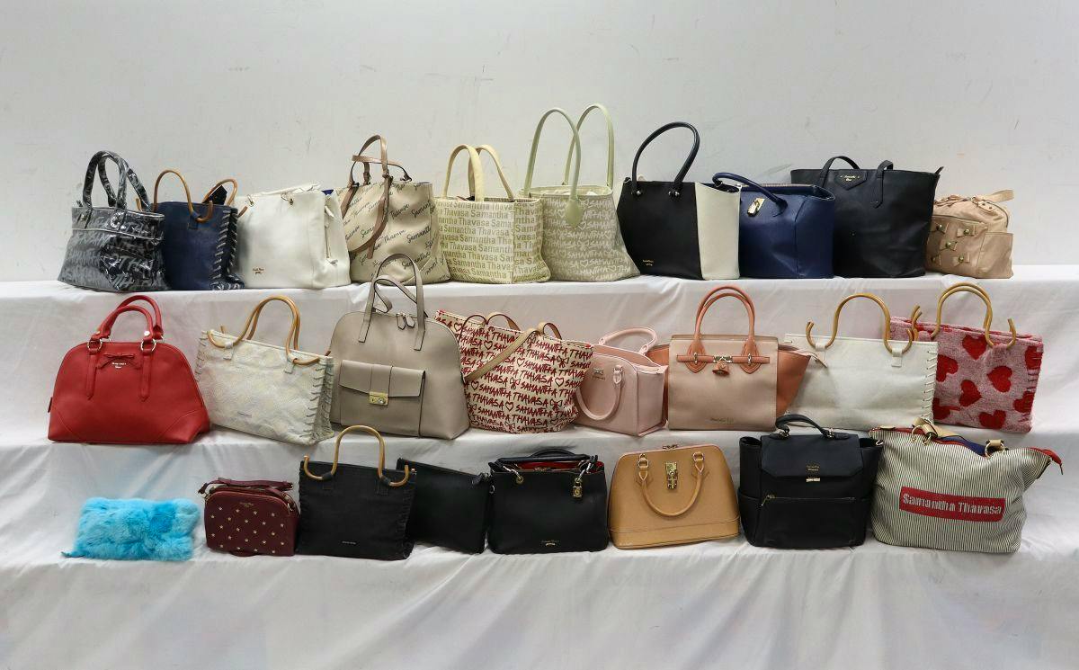 Multiple Samantha Thavasa bags displayed on three white tiers against a plain wall.