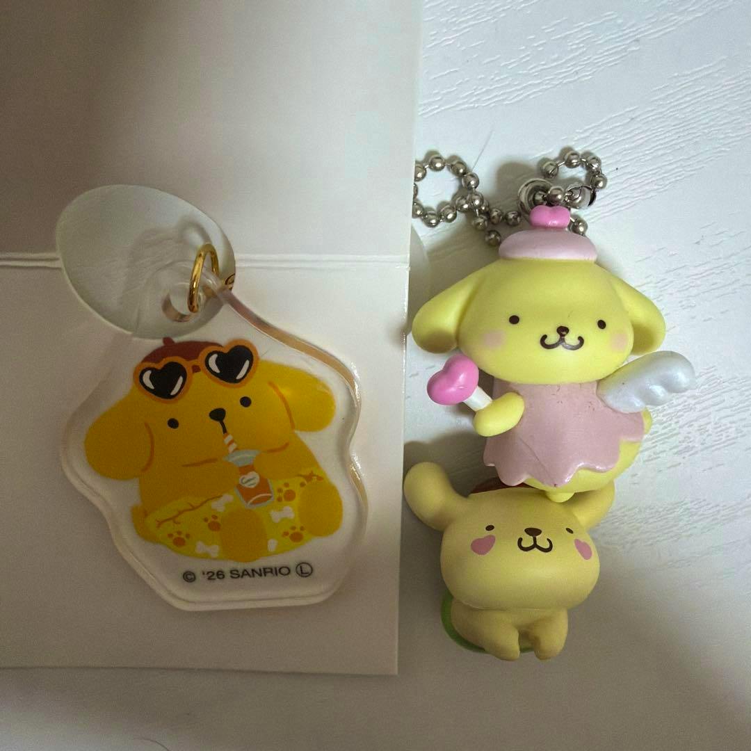 A flat acrylic Pompompurin keychain showing the character with sunglasses beside a 3D Pompompurin angel figure keychain on a ball chain.