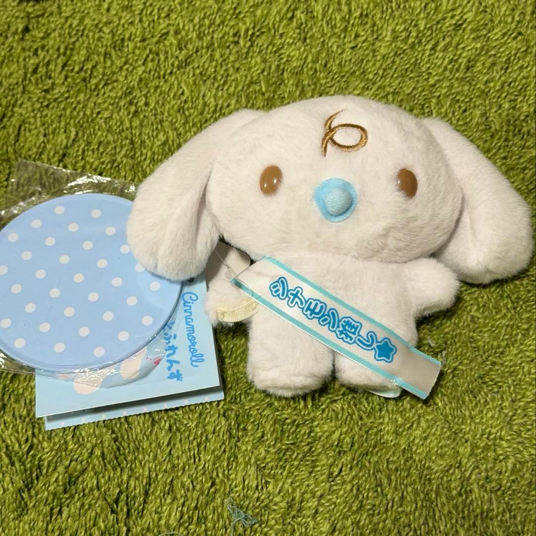 Front view of a white Cinnamoroll plush with a blue nose and a ribboned tag, shown on a green textured surface next to a blue polka-dot accessory.