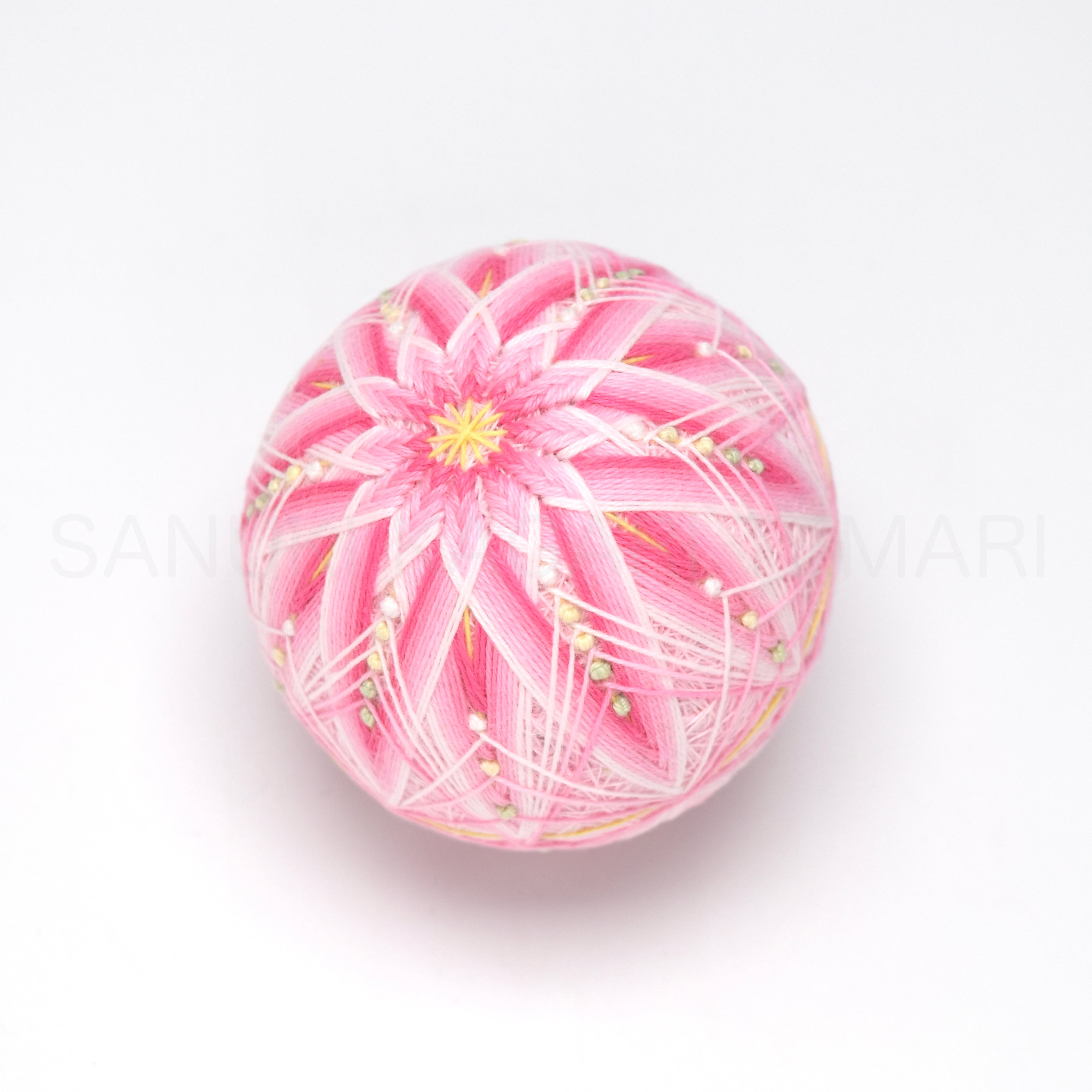 Pink embroidered fabric temari ornament shown from above with a white and yellow flower-like pattern.