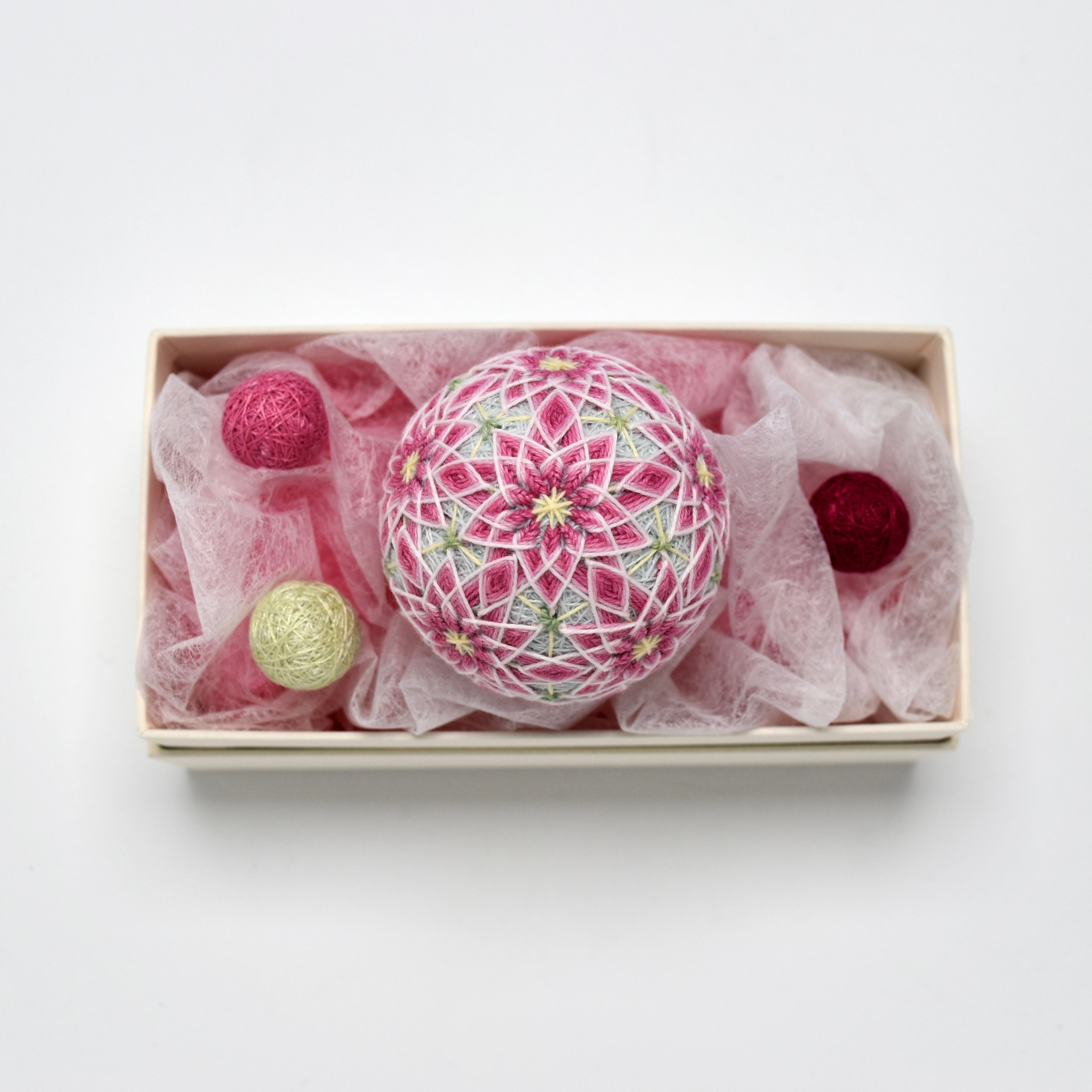 Pink and white embroidered temari ornament displayed in a rectangular box with pink tissue and three small yarn balls.