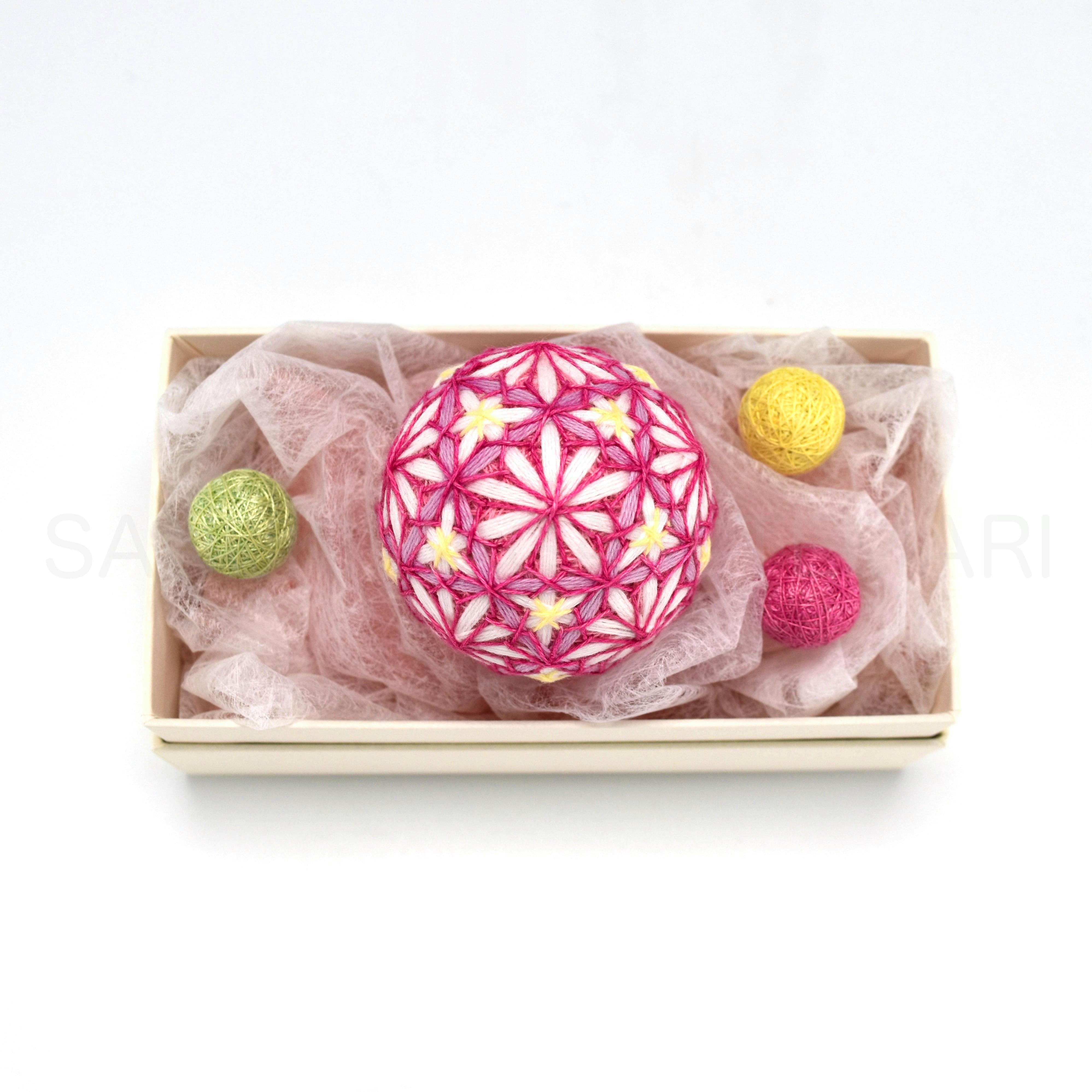 A cream-colored box holding a pink embroidered temari ball and several smaller colored yarn balls on pink tissue.