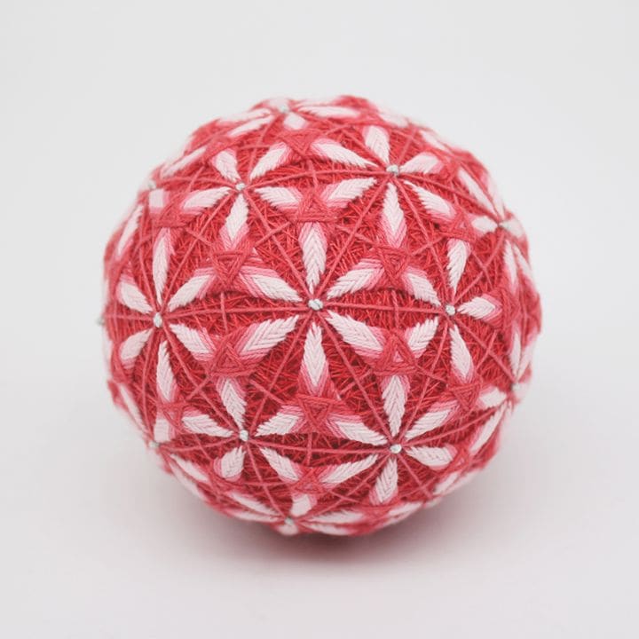 Red and white embroidered temari ornament ball with geometric flower-like stitching on a white background.
