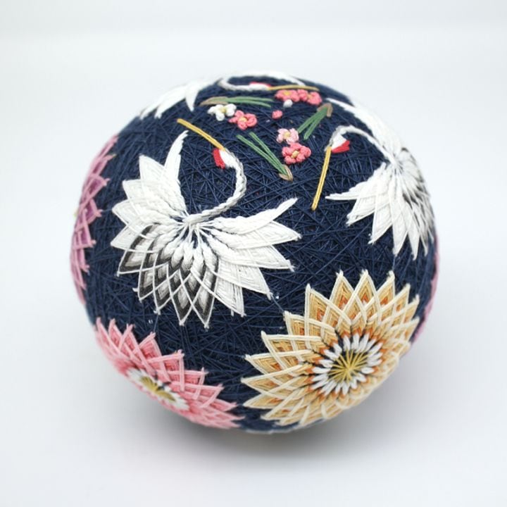 A navy embroidered temari ornament featuring white crane motifs and pink and yellow floral patterns.