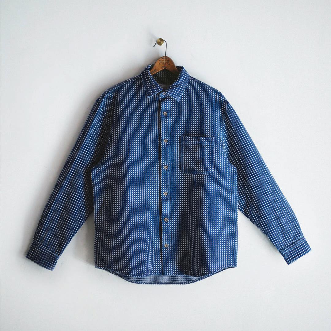 Front view of a men's indigo sashiko patterned long-sleeve cotton shirt on a wooden hanger against a white wall.