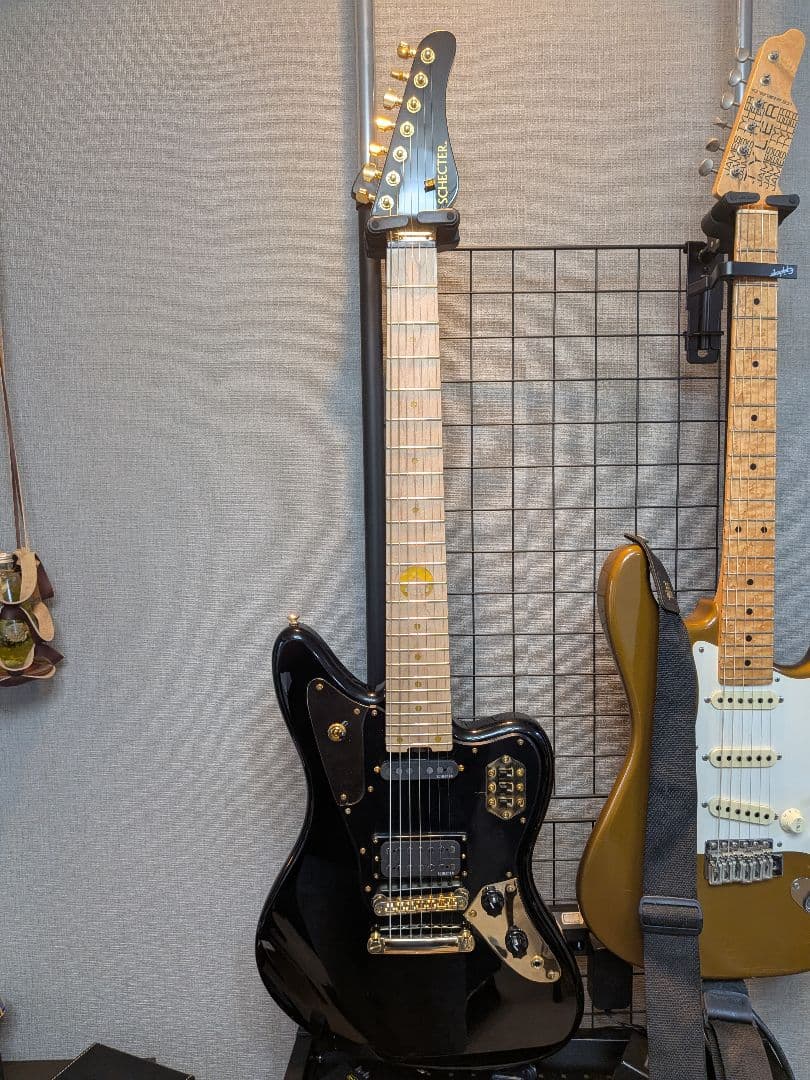 Black SCHECTER BC-AveMujica/Doloris electric guitar with a maple neck hanging on a wall grid next to another guitar.