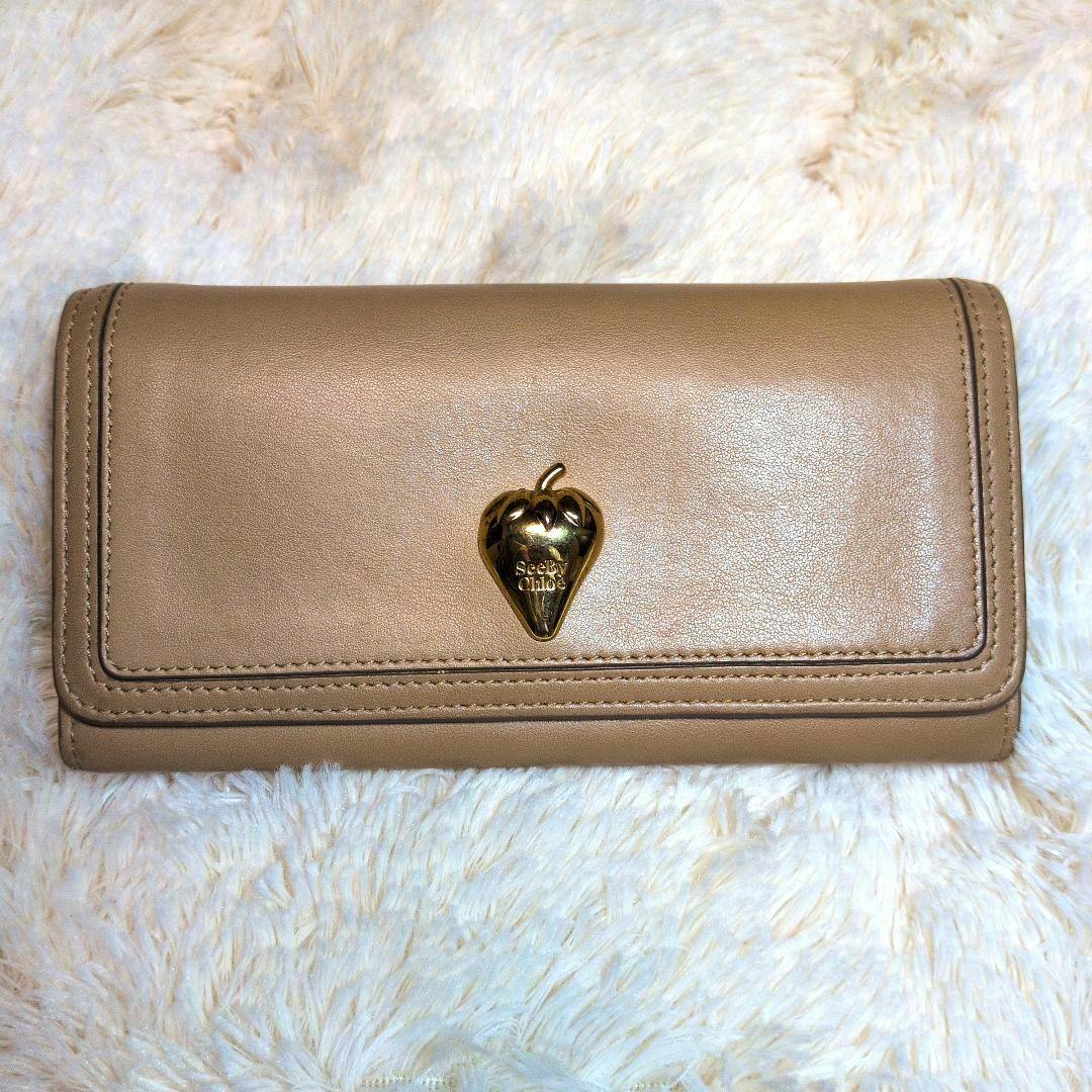 Beige SEE BY CHLOE long wallet with a gold strawberry motif placed on a light fuzzy background.