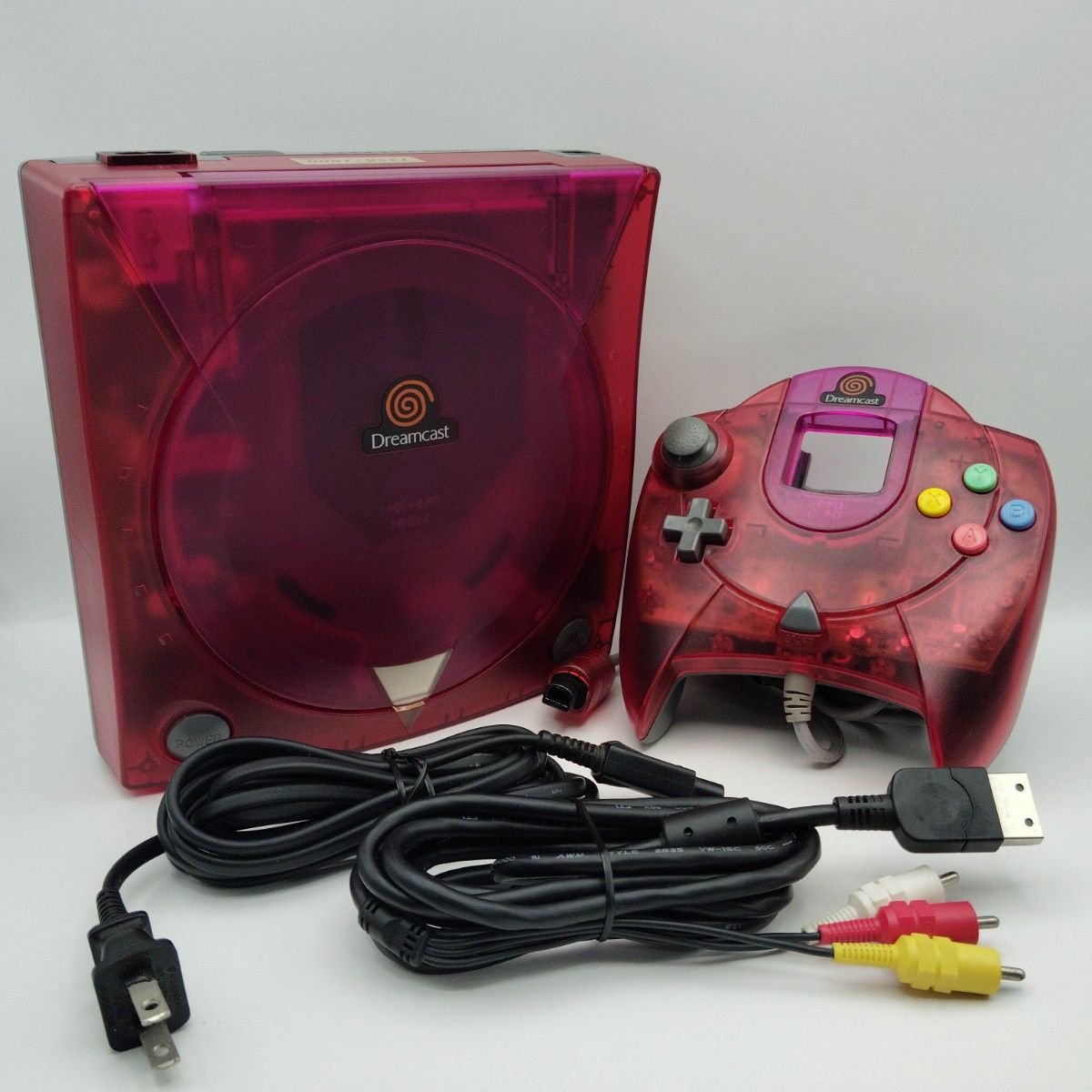 Translucent pink Dreamcast console with matching controller and AV and power cables on a white background.