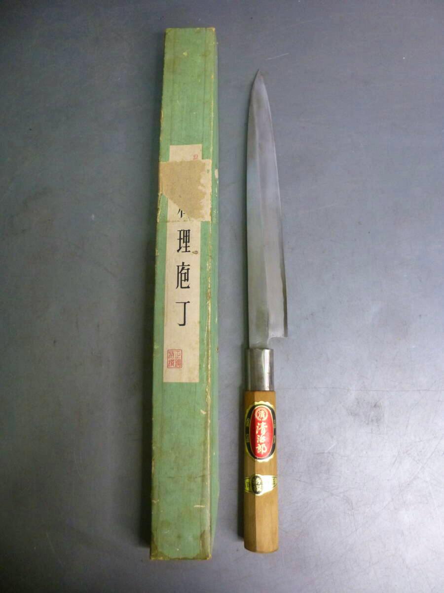 A used sashimi chef knife (blade about 23 cm) with a wooden handle and metal ferrule displayed next to its green storage box on a gray surface.