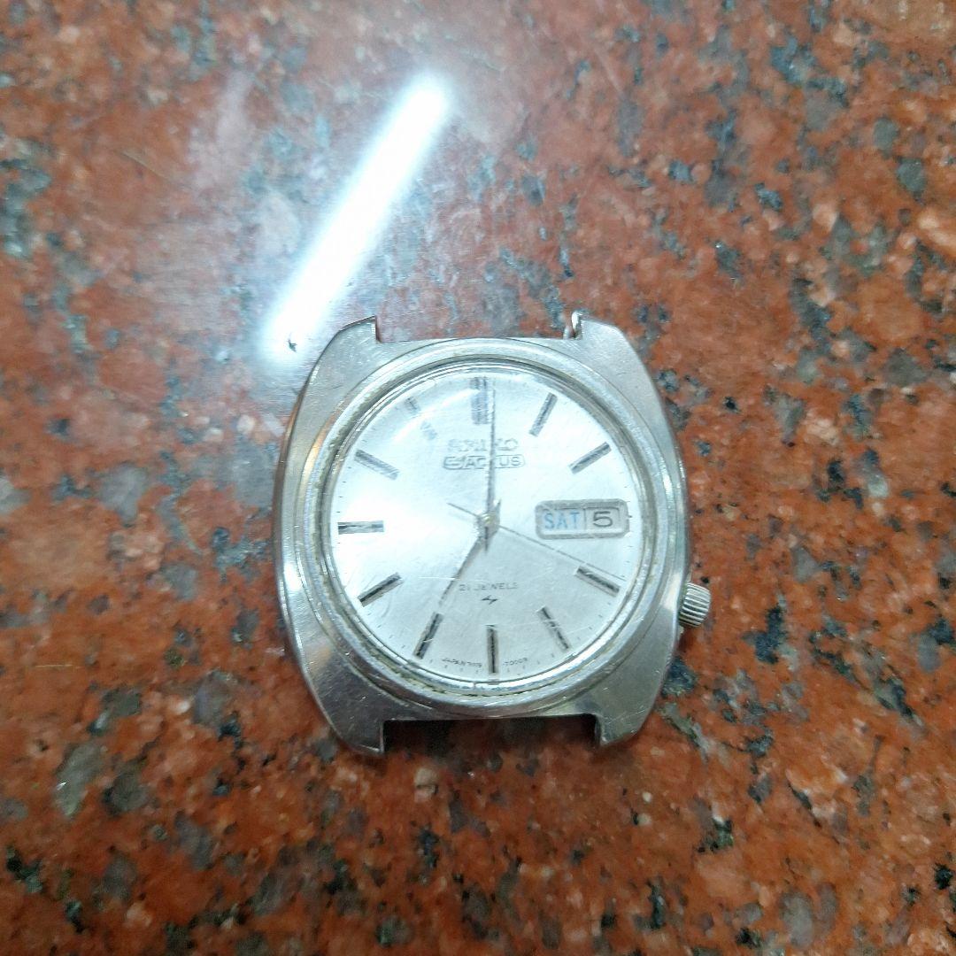 Front view of the SEIKO 5 watch case showing the dial and day-date window on a mottled stone background.
