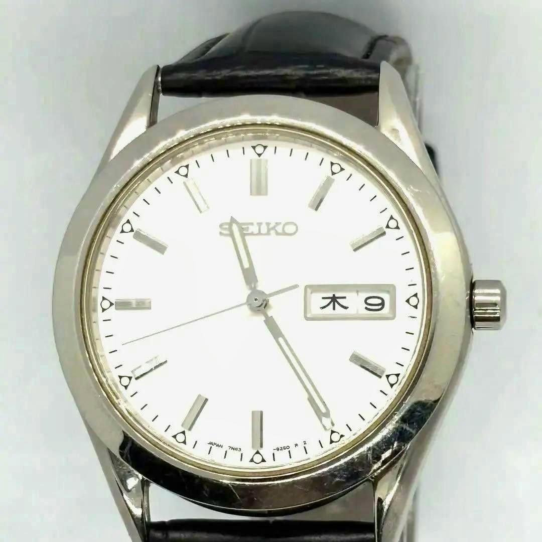 Close-up front view of a SEIKO 7N43-9080 quartz wristwatch with a white dial, day‑date window at 3 o'clock, silver metal case, and black leather strap.