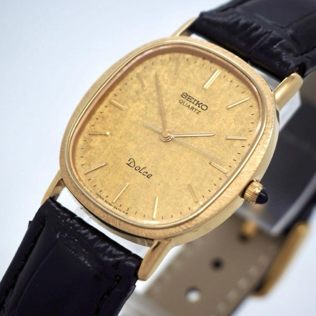 Close-up of a gold oval Seiko Dolce men's watch showing the textured dial, hands, and black leather strap.