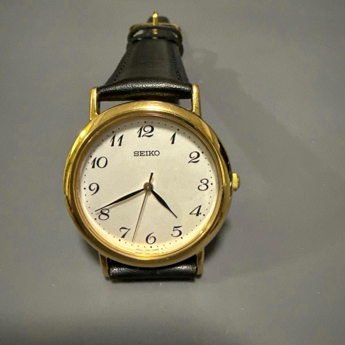 Seiko wristwatch with a gold metal case, white dial with black Arabic numerals, and a black leather strap.