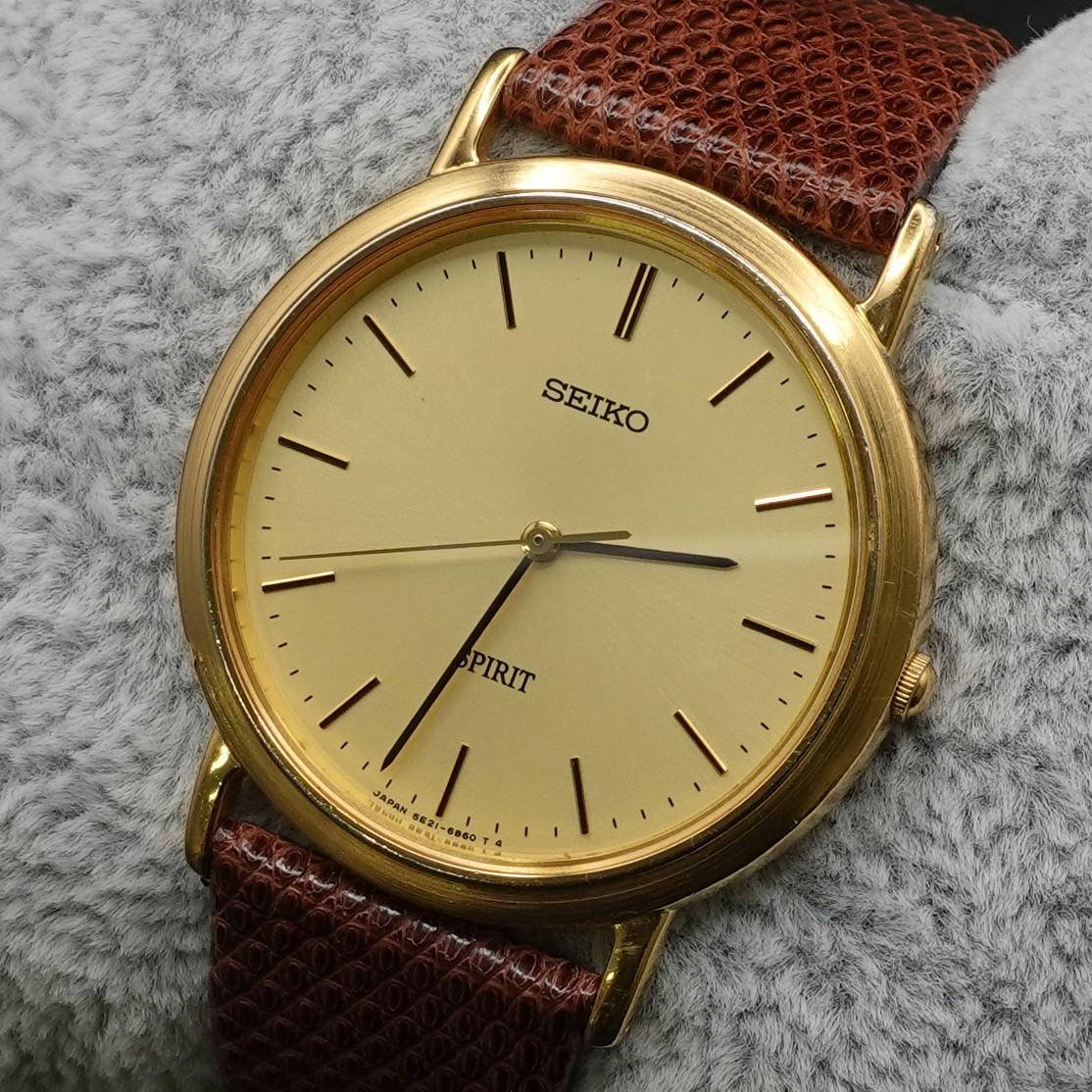 Close-up of a SEIKO SPIRIT gold dial wristwatch with a brown leather strap on gray fabric.