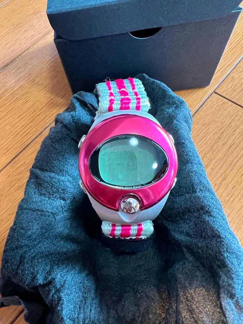A SEIKO SPOON 12-000 commemorative watch with a pink bezel and striped fabric strap resting on a black cushion with its box behind it.