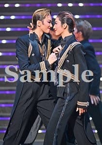 Stage photograph of two performers in dark military-style costumes embracing on a lit stage with a 'Sample' watermark.