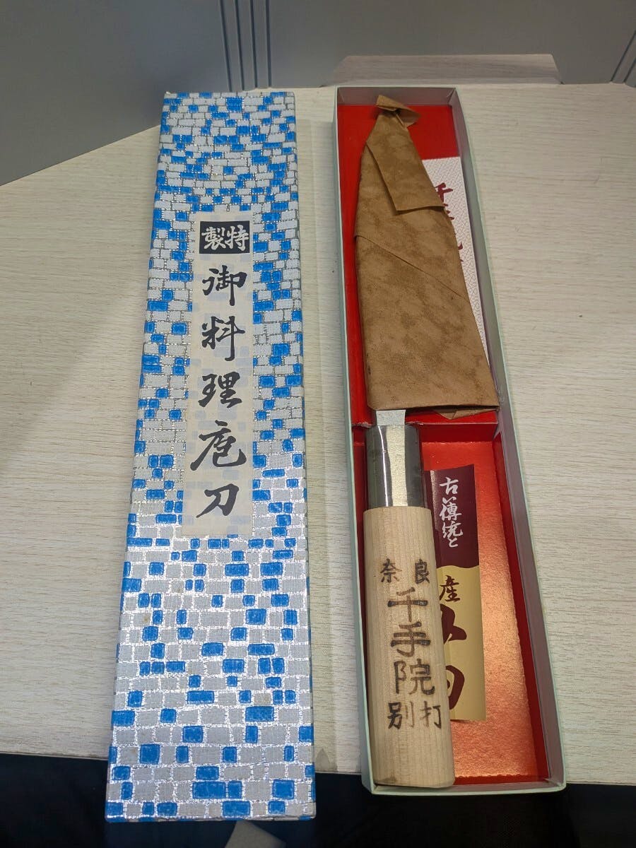 Chef knife with a wooden handle and wrapped blade displayed in its box next to a blue-and-silver patterned lid.