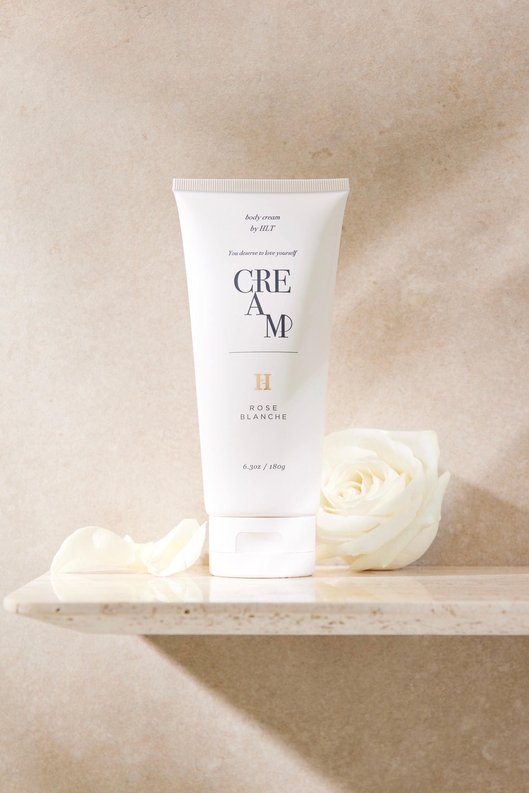 Upright tube of SENSUAL RICH BODY CREAM - ROSE BLANCHE on a stone shelf with white roses.
