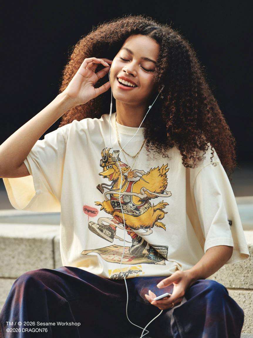 Person seated wearing a cream-colored Sesame Street Market × Dragon76 graphic T-shirt showing a large printed yellow character, listening to earphones.